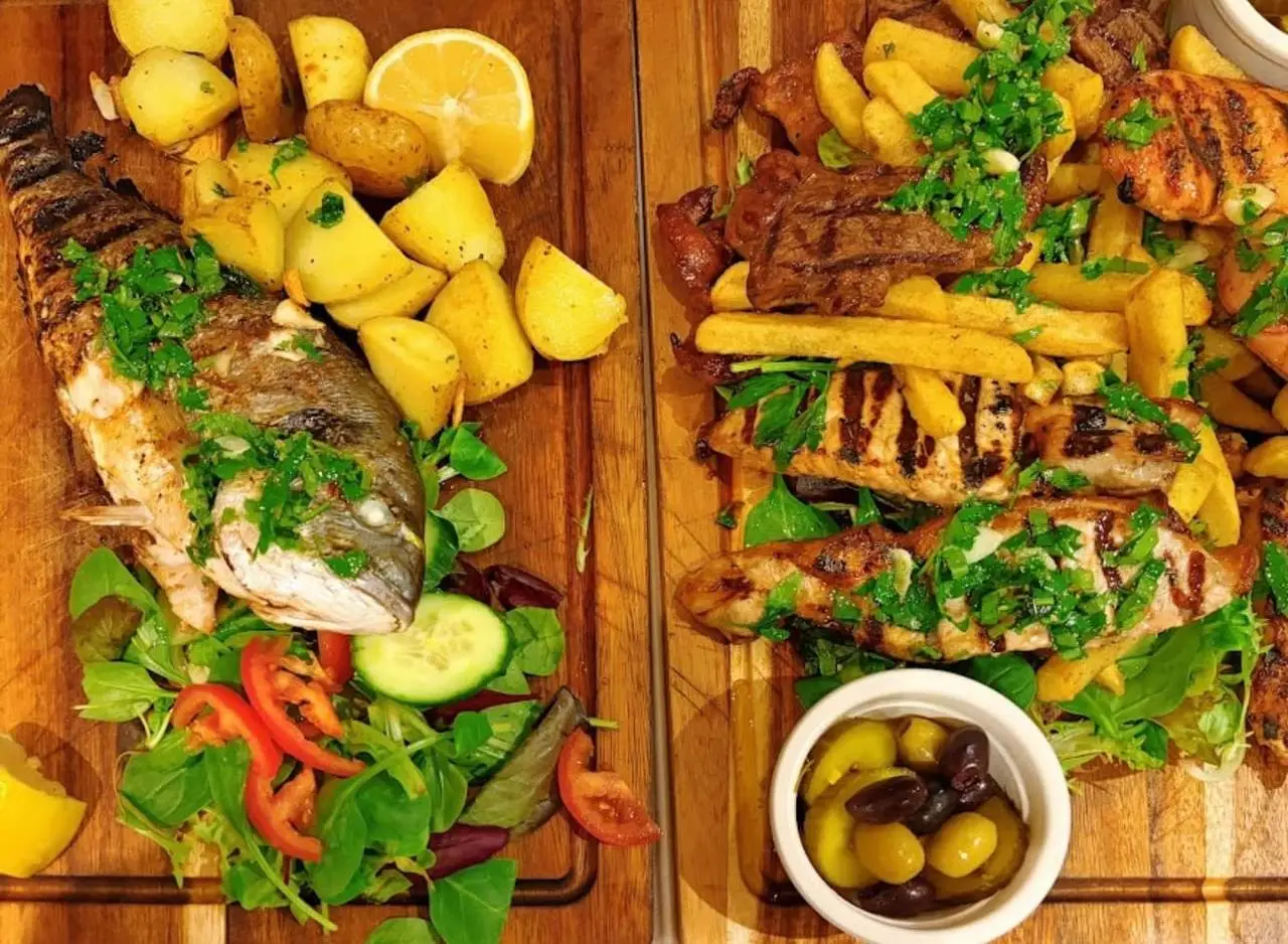 A whole grilled sea bream on a board beside a board of mixed grilled meats with chips and olives