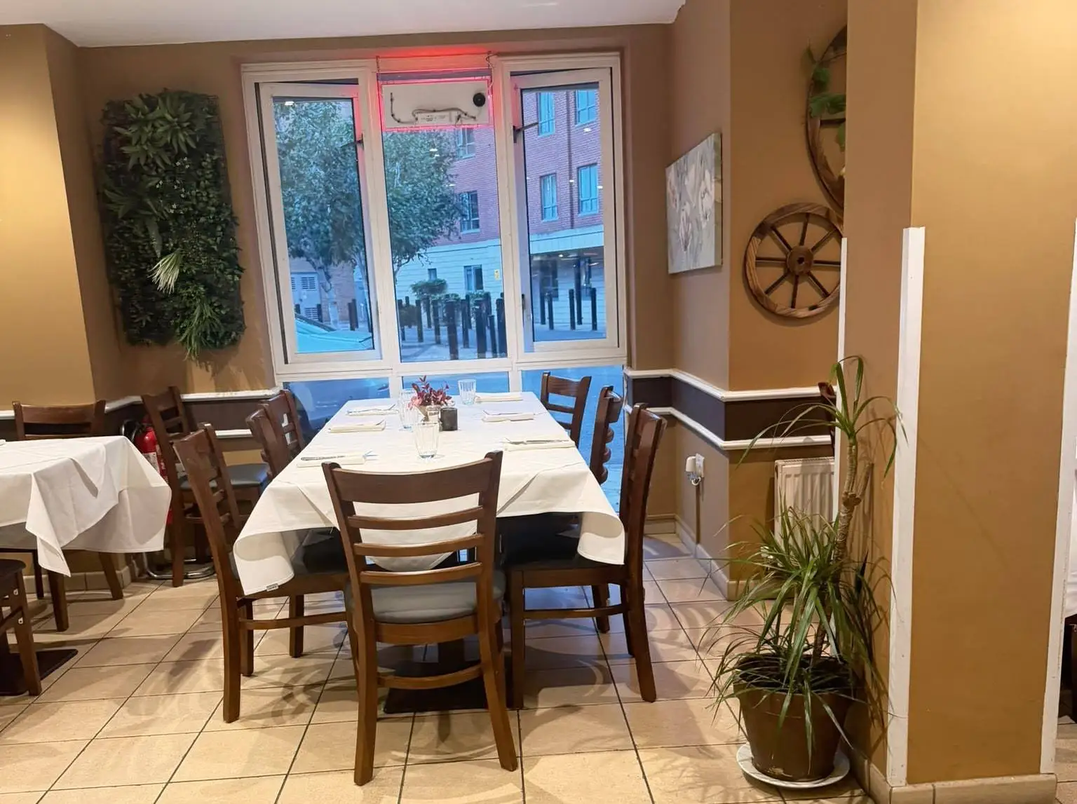 Table for eight with white cloths beside a tall window, a wooden cart wheel and a living green wall